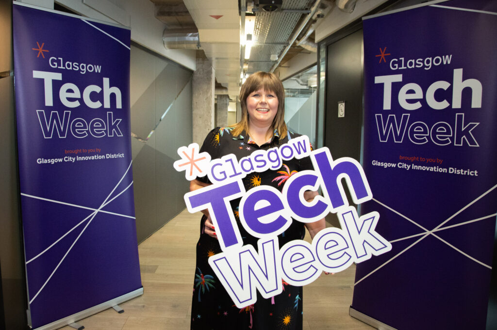 Sooz Young at Glasgow Tech Week, holding a branded sign and representing Technology Coaching as a digital skills leader. The event, hosted by Glasgow City Innovation District, highlights innovation, digital skills, and the future of technology in Scotland.